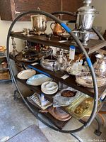 Wide view of round wood and metal shelf containing various silver tea sets, glassware, plates, and trays.