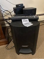 Photo showing the Bose branded subwoofer with two small speakers and a remote control on top, cables visible.