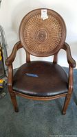 Front view of wood frame armchair showing round woven wicker back and dark brown leather seat with metal tacks on base.