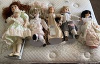 Five collector dolls arranged on a mattress: from left to right a large Seymour Mann doll with red hair in vintage pale green dress, three smaller dolls including three Brinns dolls with various hair colors and clothes, and one unmarked doll in light pink outfit.