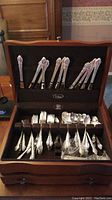 Open wooden chest displaying assorted silver plate flatware including forks, knives, and spoons arranged in slots and drawers.