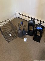 Full view of metal wire basket, metal desk lamp, and two black metal lantern-style candle holders positioned on floor near baseboard heater.