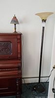 Wide view showing both lamps side by side near a wooden piano, table lamp on top of piano, floor lamp standing on floor, power cords visible.
