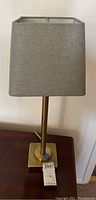 Brass tone lamp with square gray fabric shade on a wooden surface, tagged 253