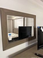 Full frontal photo of wall-mounted rectangular mirror reflecting room elements including bed and TV.