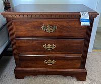 Front view of wooden nightstand with three drawers and brass handles, showing carved trim and fluted corners.