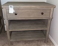Front view of the grey wooden nightstand showing single drawer with two bronze knobs and two lower shelves, on carpeted floor against wall.