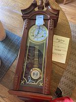 Full view of the Strausbourg Manor wooden wall clock on a table with instruction manual beside it, showing the detailed carved wood and glass front with pendulum.