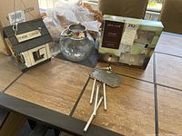 Photo of lot contents including boxed Allen & Roth string lights, wooden Penn State bird house, metal wind chime, and glass candle holder on wooden table under window light.