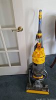 Front view of Dyson DC15 vacuum cleaner with clear dust bin and yellow handle against a white door.