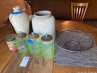 Photo of the lot showing three Vaseline glass canisters with lids, a metal wire basket, a vintage flour tin, a large white crock jar with French writing, a tall textured vase, and plaid woven placemats stacked underneath the basket.