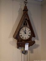Full view of the wooden wall clock with a pointed roof design, showing its overall shape, pendulum, and finial on top.