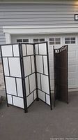 Two privacy screens standing outdoors in front of garage door; black framed one with white panels and woven wicker screen.