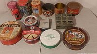 Top view of twelve assorted vintage advertising tins in various sizes and styles on a white surface showing wear and patina.