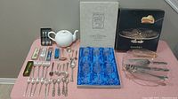 Wide view of all items including blue crystal goblets, white teapot, boxed flatware, and utensils on cake plate.