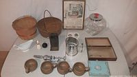 Photo of a collection of old kitchen items including a rusty cast iron pot, metal sifter, copper measuring cups, ceramic gravy boat, rotary egg beater, small box, and vintage framed kitchen cabinets advertisement by Sellers.