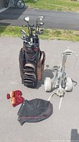 Photo showing golf bag filled with multiple golf clubs and accessories placed next to a golf cart trolley on pavement with grass in background.