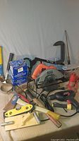 Photo showing a variety of tools including a handsaw, hammer, level, caulking gun, hydraulic jack, drill, and circular saw laid out on a surface.