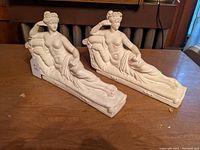 Photo of two plaster statues of partially draped reclining women on chaise lounges seen from the front and side angles on a wooden table.