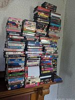 Three stacks of VHS tapes in plastic clamshell cases arranged on a wooden surface against a textured wall.