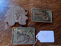Top view of three brass belt buckles on wood surface, showing Sitting Bull head, Police badge with revolver and star, and Colt Python .357 Magnum revolver designs