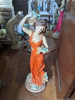 Front view showing woman in orange dress holding birds, visible paint flaking on dress and base.