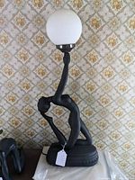 Side view of the black resin figural lamp showing the nude woman figure holding the globe light above her head, with detailed view of base and figure posture.