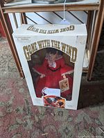 Side view of white box with 'Gone With The Wind' in gold letters and plastic window showing doll in red dress with black belt and holding a small card.