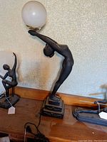 Full view of a black matte finish nude female figural lamp holding a frosted white globe lampshade in an Art Deco style.