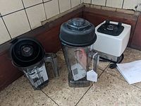 Vita-Mix blender base and two blending containers with lids placed on floor corner.