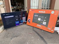 70-inch ONN Roku TV powered on with home screen menu and original retail box standing next to it