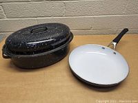 Blue enamel roaster with lid and white ceramic frying pan with black handle on a flat surface.