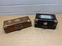 Two wooden decorative jewelry boxes side by side on a wooden surface with a brick wall background, showing overall appearance, shape, and hardware details.