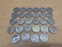 Flat lay of 28 Kennedy Half Dollar coins arranged on a brown surface in rows showing obverse side with profile of John F. Kennedy and visible dates.