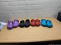 Four pairs of Crocs clogs in purple, black, red, and blue lined up side by side on a table against a cinder block wall.