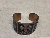 Copper bangle with etched black tribal thunderbird design on a light surface.