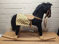 Front-left angled view showing the black rocking horse with yellow patterned saddle blanket on wooden rockers against a concrete wall background.