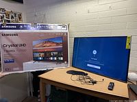 Samsung TV displayed on table powered on with no signal screen. Remote and power cable on table.
