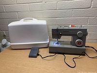 Singer Heavy Duty sewing machine with white carrying case and black foot pedal on wooden surface against brick wall background.