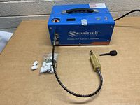 Compressor along with brass fitting connected to flexible hose and plastic bag with white and metal attached parts.