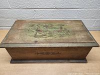 Top down view of rectangular wooden box with painted Asian landscape on lid, showing wear and fading.