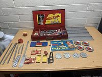 Overview photo of vintage Erector Set components arranged on table including metal beams, wheels, pulleys, plates, wooden box with label, and instruction booklets.