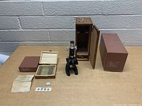 Overall view of vintage microscope with wooden storage box, wooden box of slides, paperwork and additional slide covers on table surface