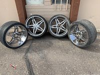 Four Lexani chrome rims with Sumitomo tires lined up against a wall outdoors, showing overall condition and size.