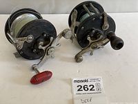 Two large metal fishing reels with handles and deep sea line spooled on, shown on white surface.
