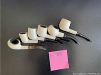 Overview of six meerschaum pipes showing cream-colored bowls and black mouthpieces, arranged side by side on black surface with a pink sticky note marked '26'.