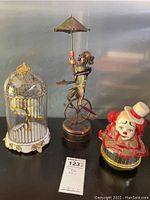 Photo of three music boxes: gilded cage with lovebirds, metal clown on unicycle with parasol, clown head with ruffled collar.
