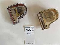 Two small piano-shaped music boxes, one with brown frame and one with cream frame, shown from top revealing internal mechanisms under clear plexiglass covers.