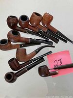 Nine used wooden smoking pipes arranged on a table, varying in bowl shape, size, and stem style. The pipes show wear and some have decorative metal inlays near the bowl or stem.