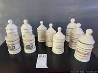 Full lot of 10 white ceramic apothecary jars with various vintage pharmaceutical logos placed on black table.